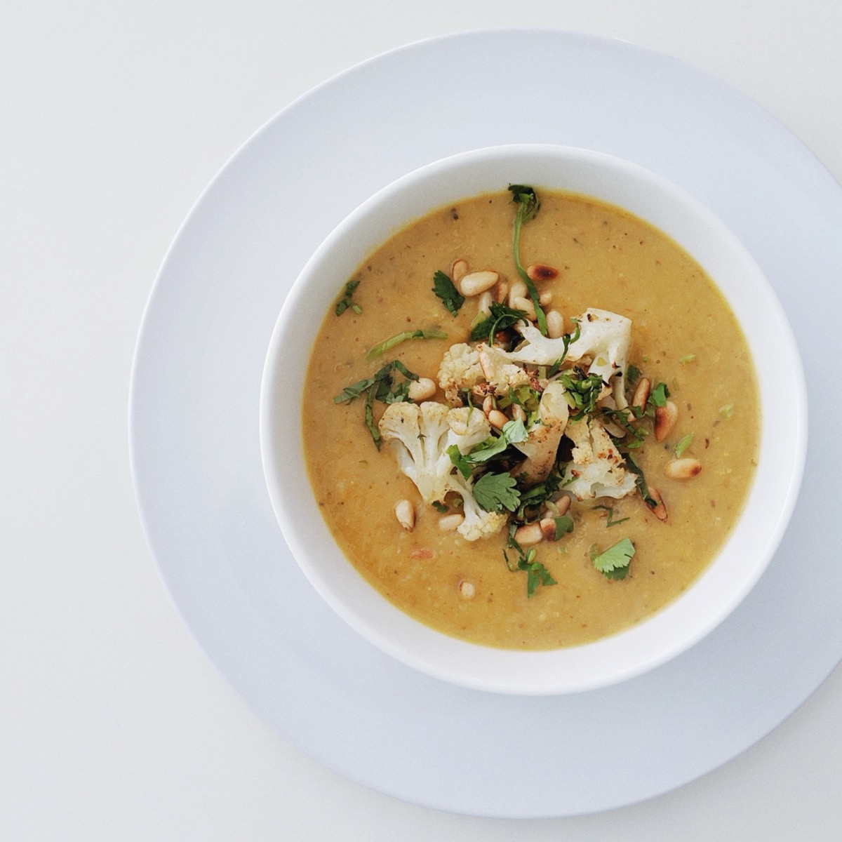 A white bowl of roasted cauliflower soup, topped with herbs and pine nuts.